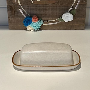 Puede incluir: Una mantequera de cerámica con tapa, en color neutro con ribete marrón. Detrás, un letrero de madera con la palabra "hello" y una corona floral de flores de fieltro en tonos azules, rosas y blancos.