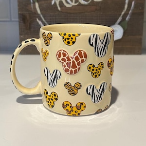 May include: A cream-colored ceramic mug with a black dotted handle and a repeating pattern of Mickey Mouse heads with animal print designs. The background features a wooden sign with the word "hello" and floral accents.