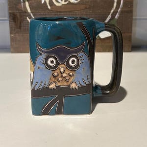 May include: A teal ceramic mug with a hand-painted owl design. The mug has a square shape with a dark handle. In the background, a wooden sign with the word "hello" and floral accents.