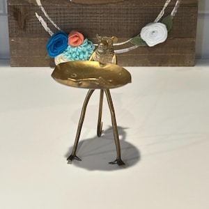 May include: A decorative wooden sign with the word "hello" in white script, surrounded by a painted white circle and colorful felt flowers. A small, gold-colored dish with a bear figurine sits in front.