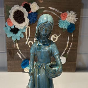May include: A teal and blue ceramic figurine of a woman in a long dress, holding a basket and a bag. The background features a wooden sign with a floral wreath. The figurine is glazed.