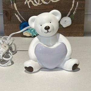 Könnte beinhalten: Weiße Keramik-Nachtlicht in Form eines Bären, der ein lila Herz hält. Der Bär hat braune Augen und eine braune Nase. Das Nachtlicht ist eingesteckt und hat ein Kabel.
