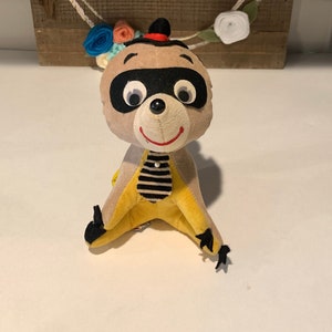 Może przedstawiać: Pluszowa zabawka w kształcie szopka z czarną maską, żółto-czarną paskowaną koszulą i czerwonym kapeluszem. Zabawka siedzi na białym tle przed drewnianym znakiem z napisem "hello".