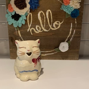 Peut inclure: Un pichet en céramique blanche en forme de chat avec un nœud rouge et une bordure bleue. Le pichet sourit et a une expression fantaisiste. Le pichet est devant une pancarte en bois avec le mot "hello" écrit en peinture blanche et une couronne de fleurs en feutre dans les tons pêche, blanc, bleu et vert.