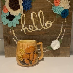May include: A brown ceramic mug with a mushroom design. The mug is sitting in front of a wooden sign with the word "hello" written on it. The sign is decorated with felt flowers in shades of blue, white, and pink.