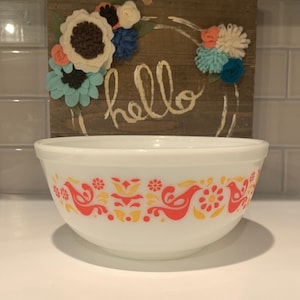 Pode incluir: Tigela de mistura de cerâmica branca com um padrão floral vermelho e amarelo com pássaros. A tigela está sobre uma bancada branca em frente a uma placa de madeira com a palavra "hello" escrita nela.