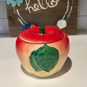Puede incluir: Un tarro de cerámica con forma de manzana, con tapa roja y tallo verde. El tarro tiene un diseño de hojas verdes. En el fondo, un letrero de madera con la palabra "hola" y una corona floral.