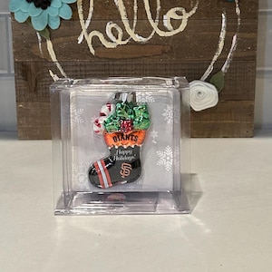 May include: A clear plastic box containing a glass Christmas ornament shaped like a stocking. The stocking is decorated with the San Francisco Giants logo and the words "Giants" and "Happy Holidays!"