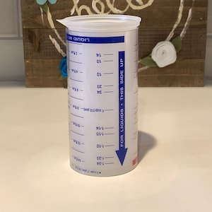 以下が含まれることがあります： 青い目盛りが付いた白いプラスチック製の計量カップ。カップには蓋が付いており、カップと液量オンスの両方が表示されています。カップは、木製の看板の前にある白い表面の上に置かれています。看板には「hello」と書かれており、青、白、ピンクのフェルトの花で飾られています。