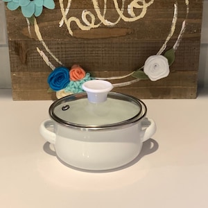 Könnte beinhalten: Ein weißer Emailletopf mit einem Glasdeckel und einem weißen Knopf. Der Topf hat zwei Griffe und steht auf einer weißen Oberfläche. Der Hintergrund ist ein Holzschild mit dem Wort "hello" in weißer Farbe geschrieben und mit Filzblumen in den Farben Blau, Rosa und Weiß verziert.