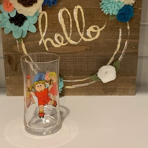 May include: A clear glass with a cartoon image of a girl with blonde hair wearing a red dress and white shoes. The girl is jumping rope with another girl. The glass has the words "Cabbage Patch Kids" printed on the bottom.