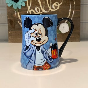 May include: A blue ceramic coffee mug with a cartoon image of Mickey Mouse wearing a blue robe and holding a red mug. Mickey Mouse has a sleepy expression and is covering one eye with his hand. The mug has a black handle.