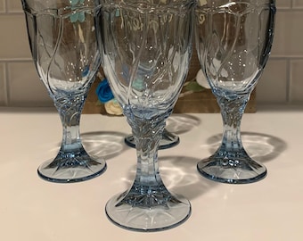 Può includere: Quattro calici da acqua con stelo azzurro chiaro con un design a foglie dettagliato. I bicchieri sono disposti su una superficie bianca, con uno sfondo in legno decorato con fiori di feltro. I calici sono alti circa 18 cm.