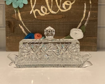 Op de afbeelding: Een glazen botervlootje van helder glas met een geslepen glasontwerp. De vloot heeft een deksel met een knop bovenop. De vloot staat op een wit oppervlak voor een houten bord met het woord "hello" in witte verf.