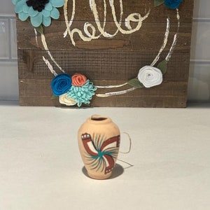 Może przedstawiać: Mały, ręcznie malowany wazon z terakoty z turkusowym, czerwonym i białym wzorem. Wazon ma pętelkowaty uchwyt i stoi na białym tle. W tle widoczny jest drewniany znak z napisem "hello" napisanym białą farbą i ozdobiony filcowymi kwiatami w odcieniach niebieskiego, białego i koralowego.