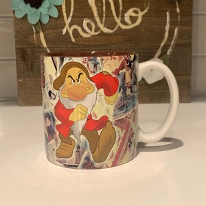Pode incluir: Uma caneca de cerâmica branca com alça apresenta uma colagem colorida de imagens do filme da Disney Branca de Neve e os Sete Anões. A caneca apresenta um close-up de Grumpy, um dos sete anões, com uma expressão rabugenta em seu rosto.