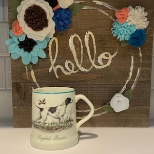 Könnte beinhalten: Cremefarbene Tasse mit einer Illustration eines English Pointers. Die Tasse hat einen grünen Rand und Henkel. Dahinter befindet sich ein Holzschild mit dem Wort "hello" und einem Kranz aus Filzblumen in verschiedenen Farben.