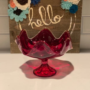 May include: A ruby red glass bowl with a scalloped edge and a pedestal base. The bowl is sitting on a white surface in front of a wooden sign with the word "hello" written on it.