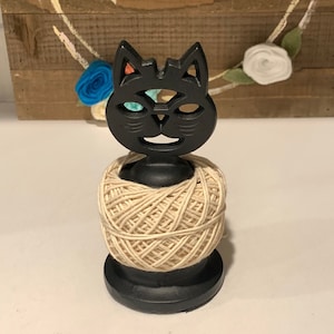 May include: Black cat yarn holder with a spool of beige yarn. The cat has a whimsical design with a multi-colored eye.