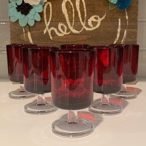 Op de afbeelding: Set van zes rode glazen met steel en heldere voetjes. De glazen zijn in een rij op een wit oppervlak gerangschikt. Het woord "hello" is in witte verf op een houten bord achter de glazen geschreven.