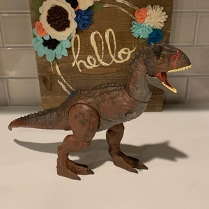 Puede incluir: Un juguete de dinosaurio marrón con marcas rojas en la cara. El dinosaurio está de pie sobre una superficie blanca frente a un letrero de madera con la palabra "hello" escrita en él. El letrero está decorado con flores de fieltro en tonos azules, blancos y naranjas.