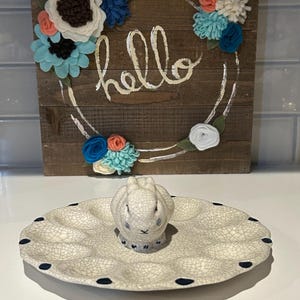 May include: A white ceramic egg plate with a bunny figurine in the center. The plate has a crackled glaze and a blue rim with black dots. The bunny is white with blue eyes and a black nose.