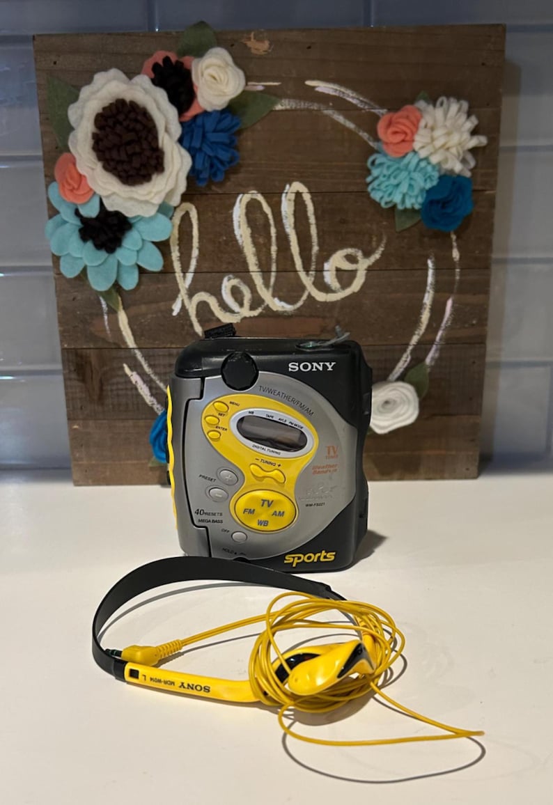 May include: A vintage Sony Sports radio with yellow accents and matching headphones. The radio has a digital display and buttons for TV, FM, AM, and WB. The word "hello" is written on a wooden background with a floral wreath.