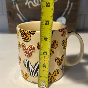 May include: A cream-colored ceramic mug with a handle, decorated with animal print Mickey Mouse head shapes. The mug is approximately 4 inches tall, as measured by a yellow tape measure. The animal prints include zebra, giraffe, and tiger patterns.