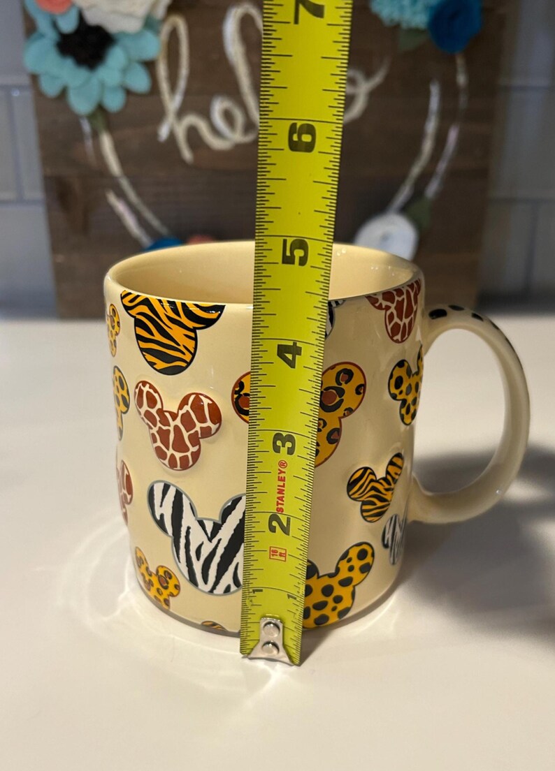 May include: A cream-colored ceramic mug with a handle, decorated with animal print Mickey Mouse head shapes. The mug is approximately 4 inches tall, as measured by a yellow tape measure. The animal prints include zebra, giraffe, and tiger patterns.