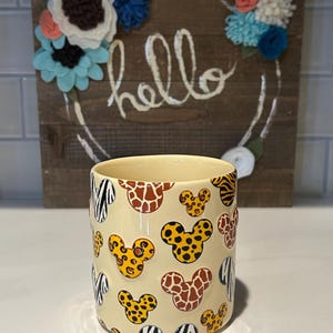 May include: A cream-colored ceramic mug with a repeating pattern of animal print Mickey Mouse heads. The background features a wooden sign with the word "hello" and felt flower accents.