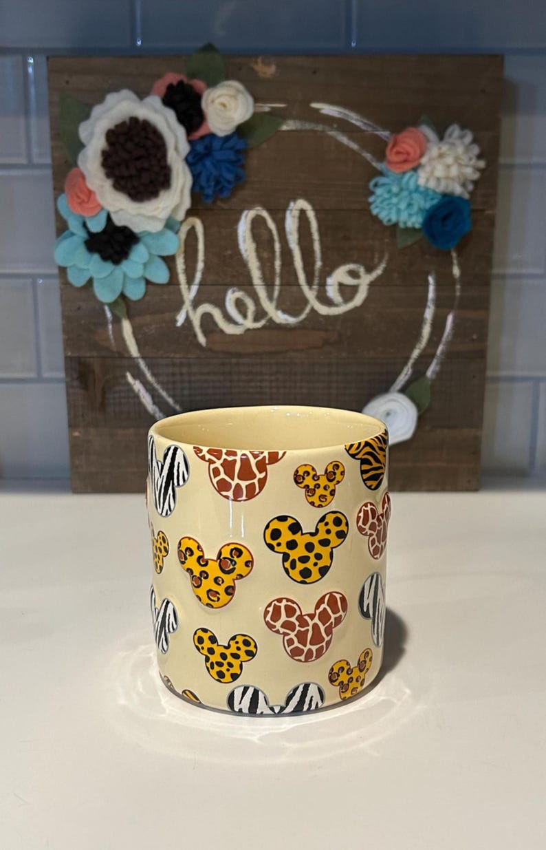May include: A cream-colored ceramic mug with a repeating pattern of animal print Mickey Mouse heads. The background features a wooden sign with the word "hello" and felt flower accents.