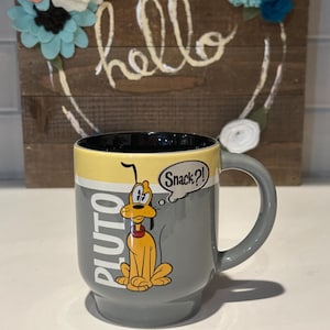 Peut inclure: Mug en céramique grise avec Pluto de Disney. L'intérieur est jaune et l'extérieur gris avec l'image de Pluto et le mot "PLUTO" en blanc. Une bulle de dialogue dit "Snack?!"
