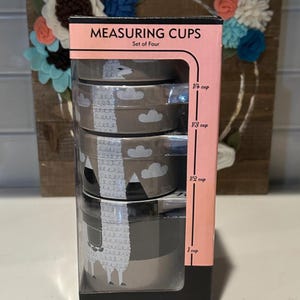 Op de afbeelding: Een set van vier maatbekers in een doos. De bekers zijn zilverkleurig met een lama- en wolkenontwerp. De doos is roze en zwart met de woorden "MEASURING CUPS" en "Set of Four". De maten zijn 1/4 cup, 1/3 cup, 1/2 cup en 1 cup.