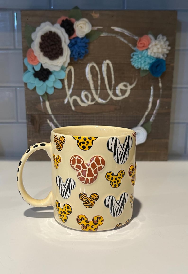 May include: A cream-colored ceramic mug with a black dotted handle and a repeating pattern of Mickey Mouse heads with animal print designs. The background features a wooden sign with the word "hello" and floral accents.