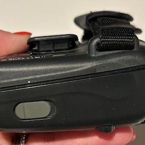 May include: A black electronic device with a black strap and a gray button. The device is held in a hand with red nail polish. The device has a curved shape and a textured surface.