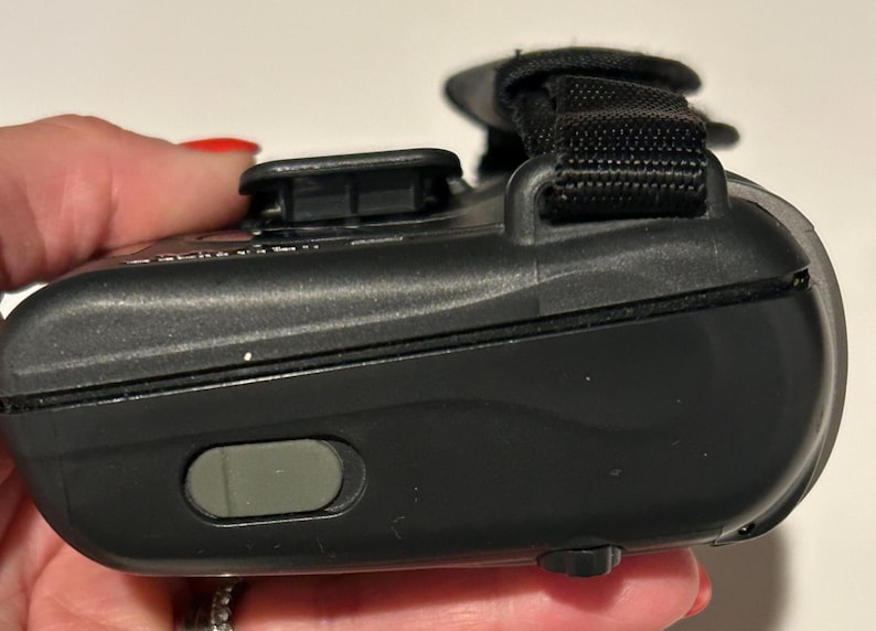 May include: A black electronic device with a black strap and a gray button. The device is held in a hand with red nail polish. The device has a curved shape and a textured surface.