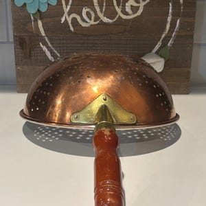 May include: A copper colander with a wooden handle and brass accents. The colander has a perforated bowl and is designed for draining liquids. The background features a wooden sign with the word "hello" and floral decorations.