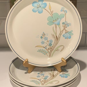 Può includere: Set di tre piatti piani in ceramica bianca con un motivo floreale blu e verde. I piatti hanno un bordo blu e sono impilati uno sopra l'altro.