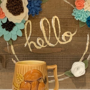 May include: A brown ceramic mug with a 3D mushroom design. The mug has a handle and is sitting on a table in front of a wooden sign with the word "hello" written on it. The sign is decorated with felt flowers in shades of blue, white, and pink.