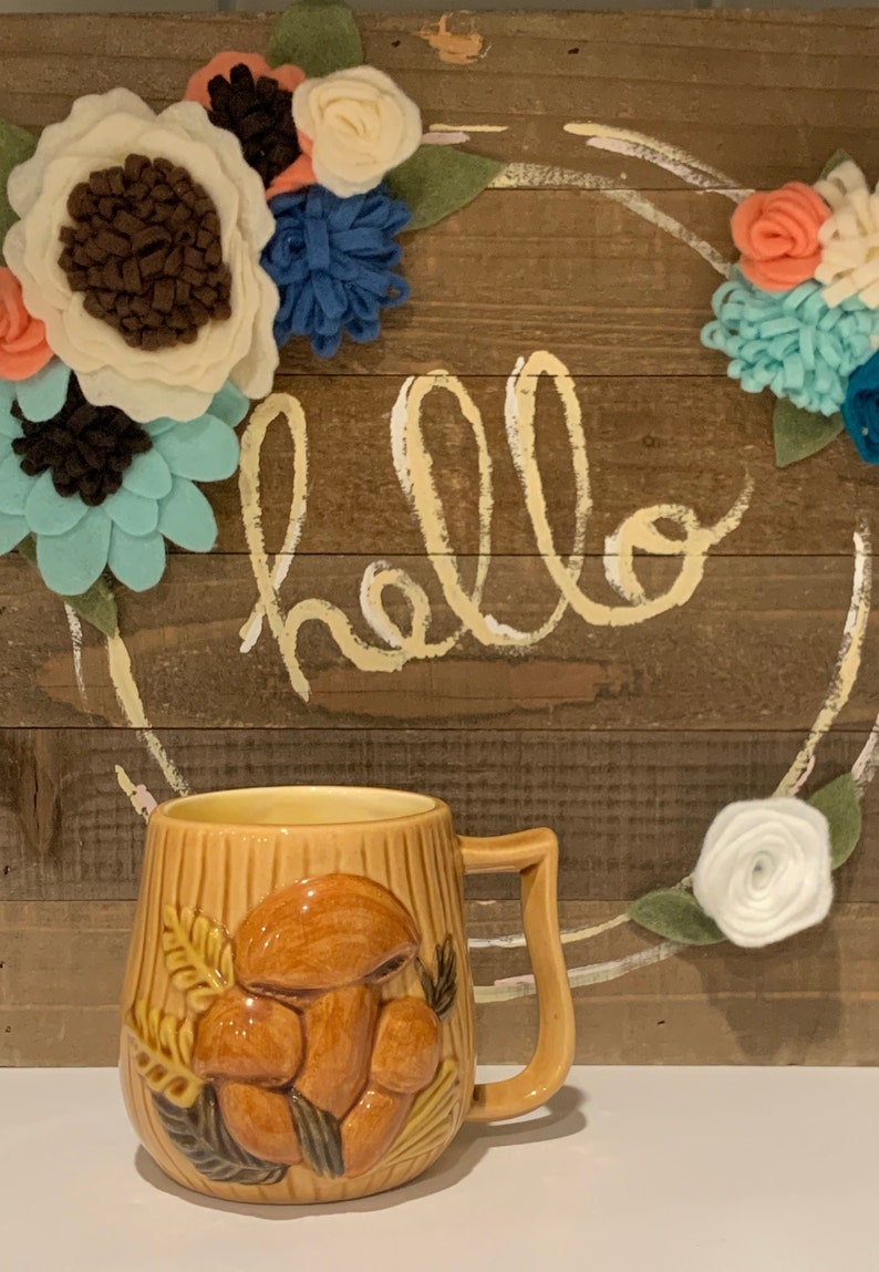 May include: A brown ceramic mug with a 3D mushroom design. The mug has a handle and is sitting on a table in front of a wooden sign with the word "hello" written on it. The sign is decorated with felt flowers in shades of blue, white, and pink.