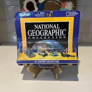 May include: A blue and yellow card with the National Geographic logo and the words "#2 Swamp Adventure" on it. The card features a small toy Land Rover, helicopter, alligator, and airboat.