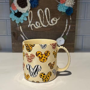 May include: A cream-colored ceramic mug featuring Mickey Mouse head shapes with various animal print patterns. The mug has a curved handle and is set against a wooden sign with the word "hello" and floral decorations.