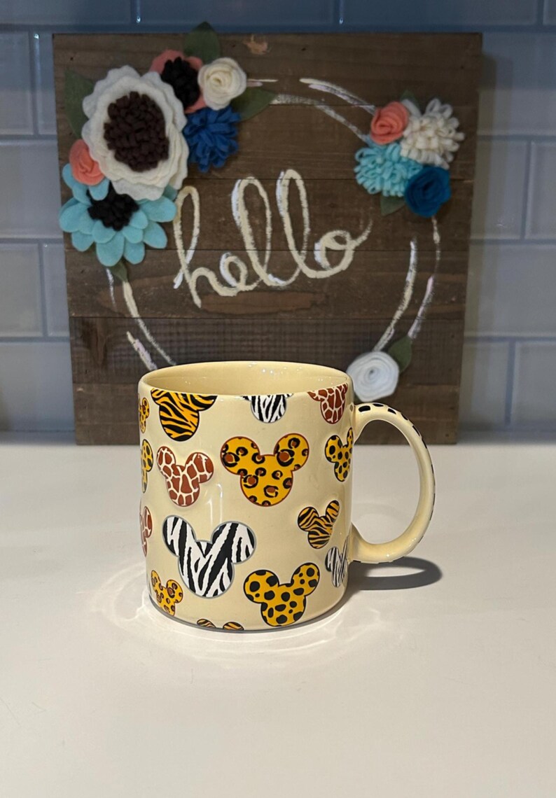 May include: A cream-colored ceramic mug featuring Mickey Mouse head shapes with various animal print patterns. The mug has a curved handle and is set against a wooden sign with the word "hello" and floral decorations.