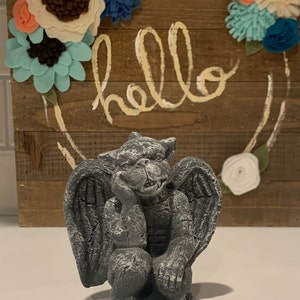 May include: A gray stone gargoyle statue with wings, sitting with its chin on its hand. The statue is in front of a wooden sign with the word "hello" written in white paint.