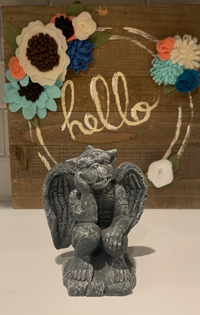 May include: A gray stone gargoyle statue with wings, sitting with its chin on its hand. The statue is in front of a wooden sign with the word "hello" written in white paint.