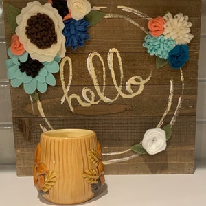 May include: A brown wooden sign with the word "hello" painted in yellow. The sign is decorated with a wreath of felt flowers in shades of pink, white, blue, and green. A brown and yellow ceramic mug with a tree design sits in front of the sign.