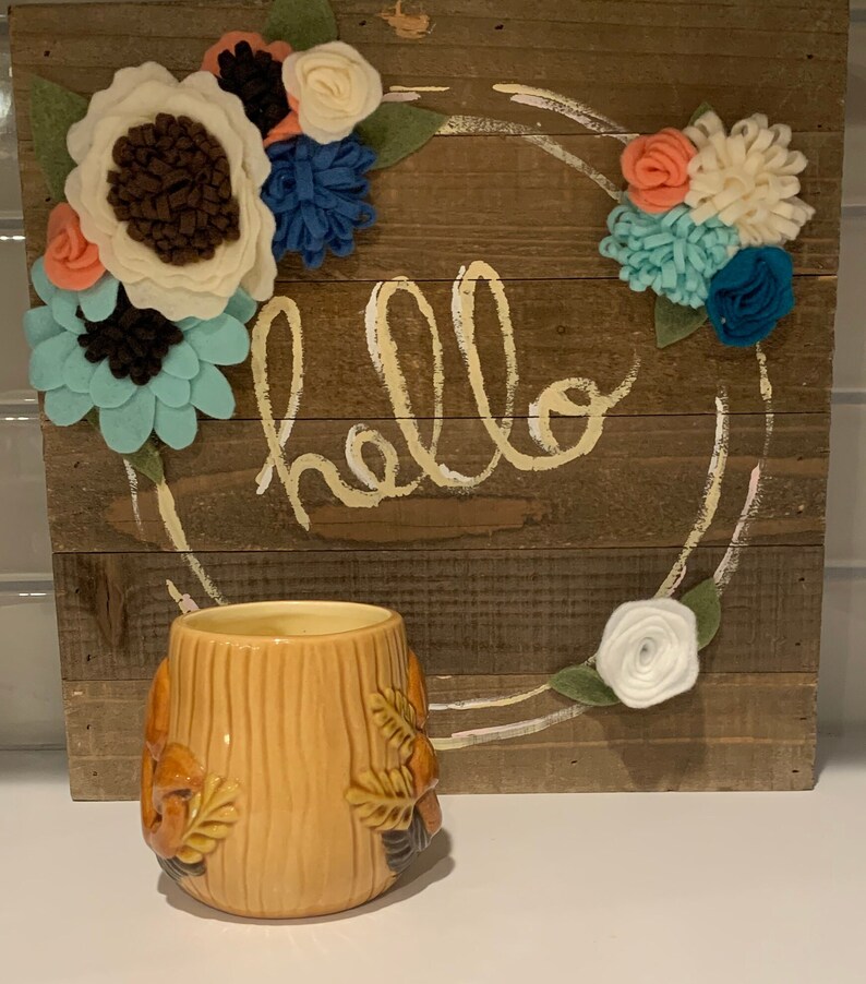 May include: A brown wooden sign with the word "hello" painted in yellow. The sign is decorated with a wreath of felt flowers in shades of pink, white, blue, and green. A brown and yellow ceramic mug with a tree design sits in front of the sign.