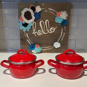 May include: Two red enamel sauce pots with silver lids and handles. The pots are on a white countertop in front of a wooden sign with the word "hello" written on it. The sign is decorated with felt flowers in shades of blue, white, and pink.