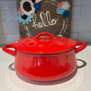 Puede incluir: Una olla de cocina de esmalte rojo brillante con tapa y asas laterales a juego. La olla está frente a un letrero de madera con la palabra "hello" y una corona floral.