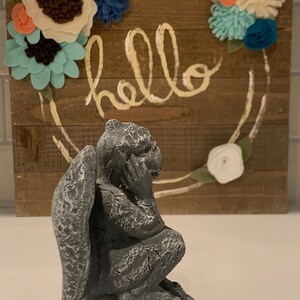 May include: A gray stone gargoyle statue with wings, sitting on a white surface. The gargoyle is looking to the right. The background is a wooden sign with the word "hello" written in yellow paint and decorated with felt flowers.